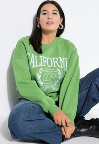 Zelená mikina s bílým textem "California" a grafikou volejbalu. Kombinována s modrými džíny a černými botami. Hladký materiál, volný střih.
