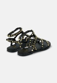 Sandalo nero con borchie a forma di piramide dorate sui cinturini, suola piatta, fibbia alla caviglia e cuciture decorative lungo i bordi.