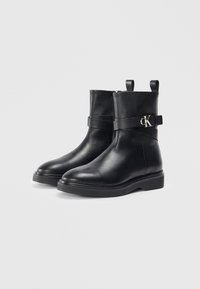 Bottines en cuir noir au fini lisse, présentant des zips latéraux, une sangle enroulante avec un logo argenté, et une semelle plate.