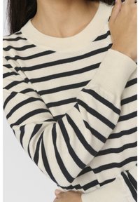 Personne portant un pull crème à rayures horizontales noires, bras croisés et gros plan sur le haut du torse et le cou.