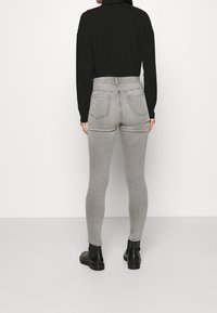 Persona vestida con jeans ajustados gris de talle alto, camiseta de manga larga negra y Botines negros, de pie frente a una pared blanca.