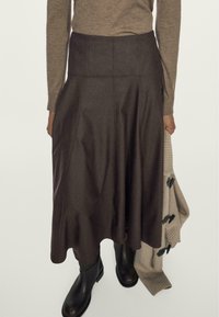 Una gonna marrone, lunga fino al ginocchio, realizzata in un tessuto liscio con un orlo svasato e ondulato, indossata con un maglione beige e stivali di pelle nera.