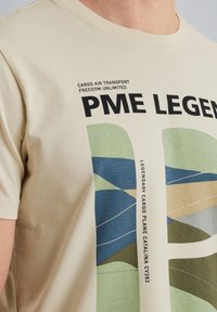 Cremefarvet bomulds-t-shirt med et grafisk design, der har blå, grønne og beige abstrakte former samt sorte tekstaccenter.