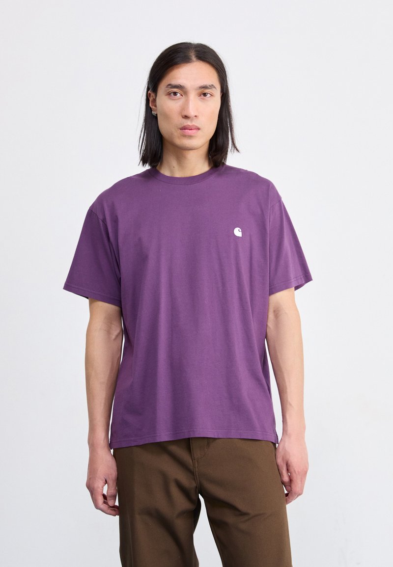 Paarse katoenen t-shirt met korte mouwen, ronde halslijn en een klein wit logo op de linker borst. Het model heeft het gecombineerd met een paarse broek.