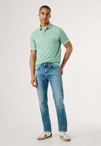Camisa polo verde claro con detalles blancos, combinada con jeans azules y zapatillas blancas. La camisa tiene un cuello y un logo en el pecho.