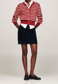 Persona vestida con un cárdigan abotonado de rayas rojas y blancas, una camisa blanca, una falda azul oscura y mocasines negros, de pie con las manos detrás de la espalda.