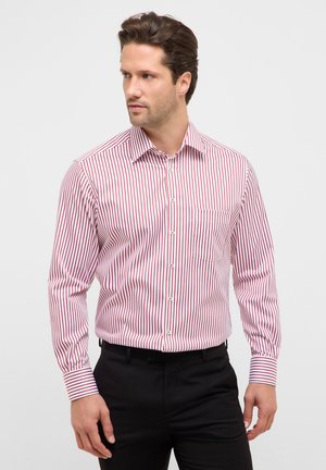 Uomo in piedi rivolto verso sinistra, indossa una camicia a maniche lunghe a righe verticali rosse e bianche e pantaloni neri, su sfondo neutro.