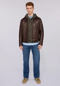 Brązowa skórzana kurtka z zamkiem błyskawicznym i bocznymi kieszeniami, noszona na zielonej bluzie z kapturem i jasnoniebieskiej koszuli, zestawiona z niebieskimi dżinsami i beżowymi trampkami.