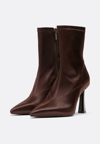Ein Paar dunkelbraune, spitze Stiefeletten mit seitlichen Reißverschlüssen und hohen, glänzenden Blockabsätzen vor einem neutralen Hintergrund.
