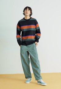 Langærmet sort sweatshirt med orange, teal og blå striber, parret med løstsiddende lysegrønne bukser og grå sneakers på et gult gulv.