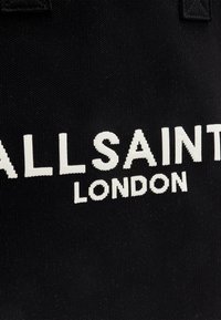 Musta kankainen olkalaukku, jossa on valkoinen brodeerattu logo "ALLSAINTS LONDON" näkyvästi esillä. Vankka kangas, minimalistinen muotoilu.