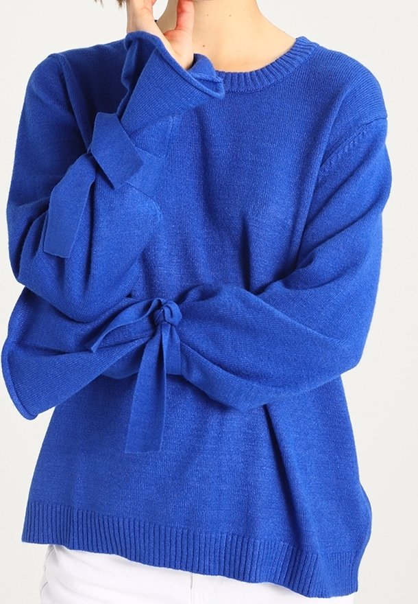Pull tricot bleu à manches longues nouées en nœuds aux poignets, porté avec un pantalon blanc, couvrant le haut du torse et les bras.