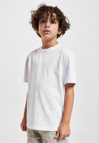Kille med lockigt hår som bär en enkel vit T-shirt och beiga byxor, stående mot en neutral ljus bakgrund.
