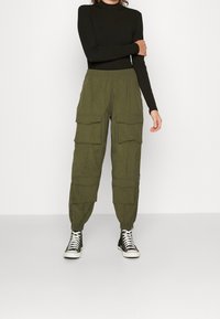 Olivgröna cargobyxor med flera stora fickor, elastiska manschetter och lätt material, i kombination med svarta högskor.