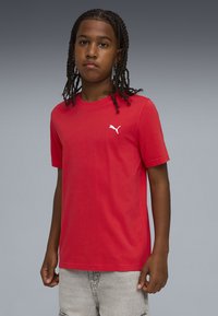 Νεαρό άτομο με πλεγμένα μαλλιά που φοράει κόκκινο t-shirt της Puma και ανοιχτό γκρι σορτς, στέκεται μπροστά σε απλό γκρι φόντο.