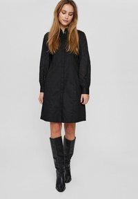 Mustmust hall dress pika pikkade tekstuuriga varrukatega, millel on lõikeavad. Sile kangas, põlvekõrgune, koos põlve kõrguste mustade saapatega.