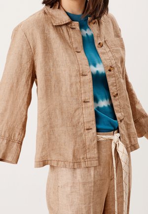 Person iført en lysebrun knappet jakke og matchende bukser med et hvidt reb-bælte over en blå tie-dye skjorte.