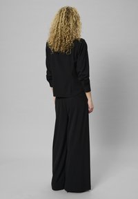 Platas kāju melnas bikses apvienotas ar atbilstošu melnu topu. Topam ir garas piedurknes, brīvs piegriezums, mīksta auduma tekstūra un minimāla apdare.