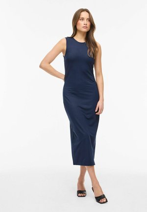 Naine seisab ühe käe puusal, kannab õlgadeta tumesinist midi kleiti ja musti avatud varbaga kontsakingi valgel taustal.