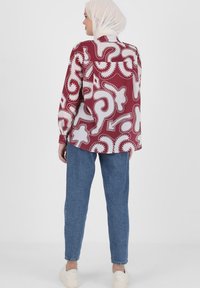 Bordeauxfarbenes Musterhemd mit weißen abstrakten Designs, lockere Passform, lange Ärmel, kombiniert mit blauen Jeans und weißen Sneakern.