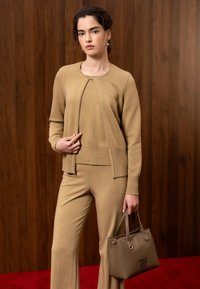 Jeune femme en tenue en tricot beige tenant un sac à main taupe, debout contre un mur en bois et un tapis rouge.