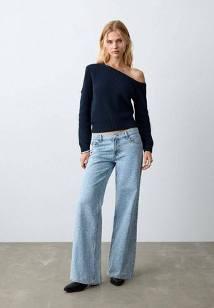 Teget pulover rebrastog uzorka s otvorenim ramenima uparен s svijetloplavim trapericama širokih nogavica s suptilnim uzorkom i crnom obućom.