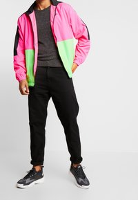 Man i en rosa och grön zip-up jacka, svart prickig skjorta, svarta uppvikt byxor och svart och grå sneakers mot en vit bakgrund.