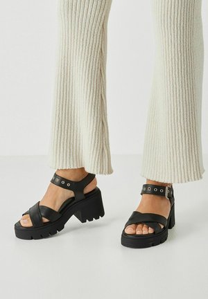 Pieds portant des sandales à talons carrés noirs avec des brides autour de la cheville, associées à un pantalon en maille côtelée de couleur crème, sur un fond blanc.