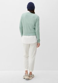 Maglione verde chiaro a trama con un taglio corto, indossato sopra una camicia bianca, abbinato a pantaloni color crema a coste e sneakers beige.