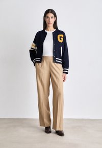 Σκούρο μπλε ζακέτα με λευκές και κίτρινες ρίγες, κεντημένο λογότυπο 'G', συνδυασμένη με μπεζ παντελόνι σε φαρδιά γραμμή και καφέ ψηλές μπότες.