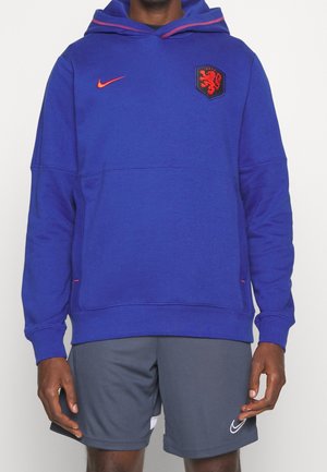 Uomo che indossa un hoodie blu della Nike con un emblema arancione di leone e pantaloni corti grigi della Nike, in piedi con le mani rilassate ai lati.