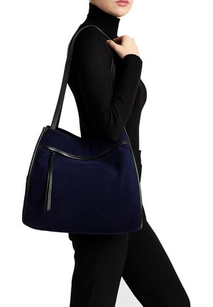 Marineblauwe suede hobo tas met een afneembare zwart leren schouderband, voorzien van een ritsluiting en een zak aan de voorkant met een strakke zwarte rand.