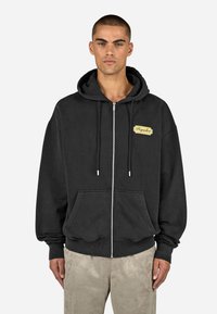 Juodas zip-up džemperis, pagamintas iš minkštos medžiagos, su priekiu kišenė, raišteliais ir kreminės spalvos etikete su užrašu "Pegador".