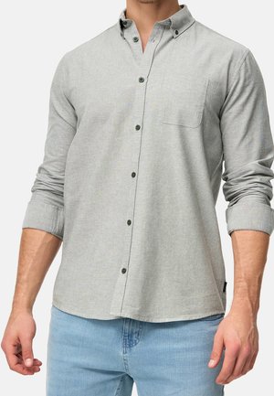 Mann trägt ein hellgraues, langärmeliges Hemd mit Brusttasche und blaue Jeans, Ärmel hochgekrempelt bis zu den Unterarmen.