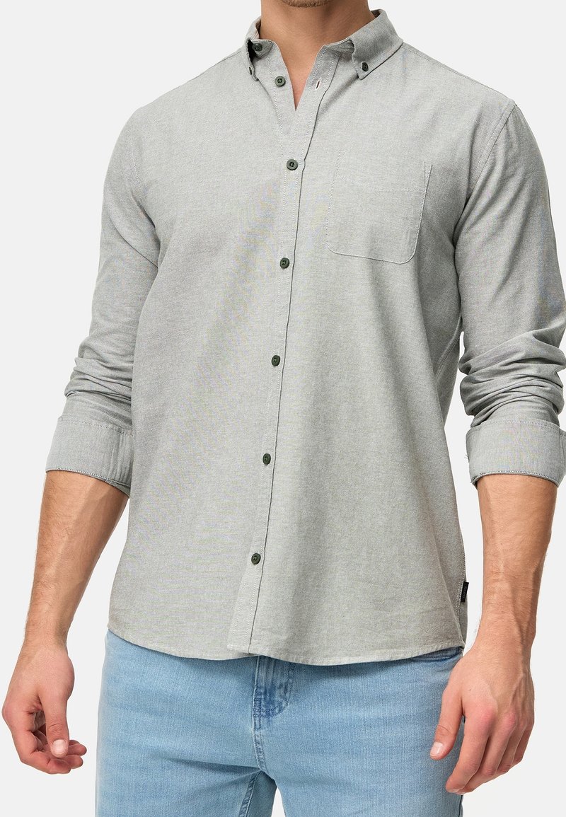 Homme portant une chemise boutonnée à manches longues gris clair avec poche poitrine et un jean bleu, manches retroussées jusqu'aux avant-bras.