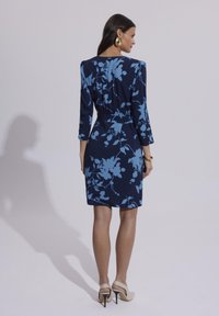 Granatowa sukienka z niebieskimi kwiatowymi wzorami, z rękawami o długości trzy czwarte i dopasowanym krojem. Modelka nosi beżowe sandały na wysokim obcasie.