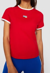 Camiseta de algodón roja con cuello y puños en blanco. Presenta un pequeño parche de logo azul y amarillo en el pecho. Manga corta, con forma ajustada.