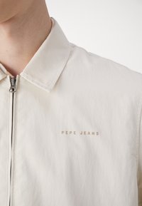 Šviesiai smėlinės spalvos striukė su užtrauktuku priekyje, pasižyminti klasikinio tipo apykakle ir minkšta tekstūra. „PEPE JEANS“ logotipas subtiliai smėlio spalvos kairėje krūtinės pusėje.