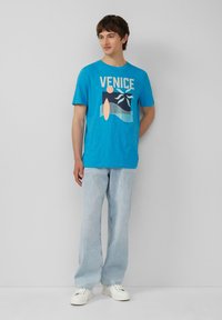 Türkisfarbenes T-Shirt mit "VENICE" Grafik, kurzen Ärmeln, Rundhalsausschnitt, kombiniert mit hellblauen Jeans und weißen Sneakers.