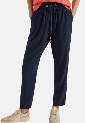 Personne portant un pantalon bleu marine à cordon, un haut corail et des baskets beiges, debout les mains dans les poches sur fond blanc.