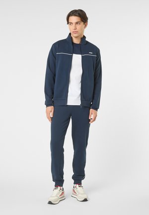 Ung mand stående iført marineblå lynlåstrøje, matchende bukser, hvid t-shirt og hvide FILA-sneakers på ensfarvet baggrund.