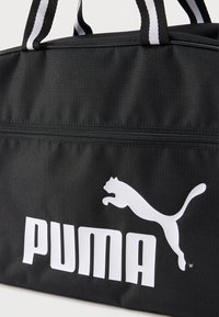 Μαύρη αθλητική τσάντα με λευκό λογότυπο Puma και όνομα μάρκας, τσέπη με φερμουάρ και χερούλια με μαύρες και λευκές ρίγες.