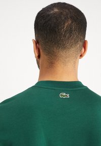 Grön bomulls-t-shirt med ribbad rund halsringning, med en liten broderad krokodillogotyp på övre ryggen. Mjuk textur.