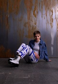 Botas de plataforma blancas con cordones, pantalones azul y blanco con patrones, y una chaqueta de mezclilla. El sujeto está sentado contra un fondo de metal texturizado.
