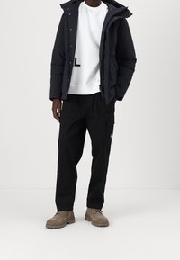Mand iført en sort hættejakke, en hvid sweatshirt med delvis sort tekst, sorte cargo bukser og beige ruskindsstøvler, der står mod en hvid baggrund.
