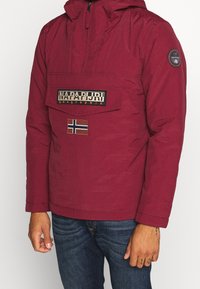 Burgundowa kurtka z kapturem z naszywką logo i norweską flagą, z kieszenią z przodu i elastycznymi mankietami, w połączeniu z dżinsowymi spodniami.