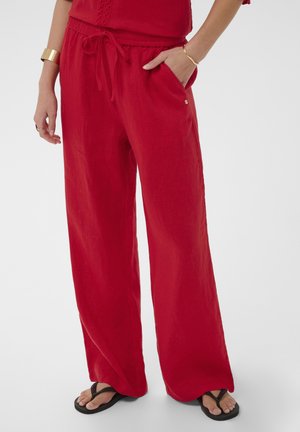 Žena nosi voľné červené nohavice s nastaviteľným pásom na šnúrku, ruku v vrecku, zlaté náramky a čierne žabky na neutrálnom pozadí.