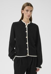 Cardigan noir avec une finition texturée, bord blanc et gros boutons. Présente un col arrondi et des manches légèrement bouffantes.