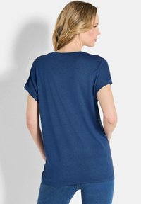 Vrouw met golvend blond haar, gekleed in een marineblauwe shirt met korte mouwen en blauwe spijkerbroek, met de rug naar de camera gekeerd tegen een effen lichte achtergrond.