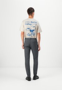 Beige t-skjorte med blå grafisk design, grå bukser og svarte sko. T-skjorten har teksten "SOHO Social Coffee Club" og tegneseriefigurer.